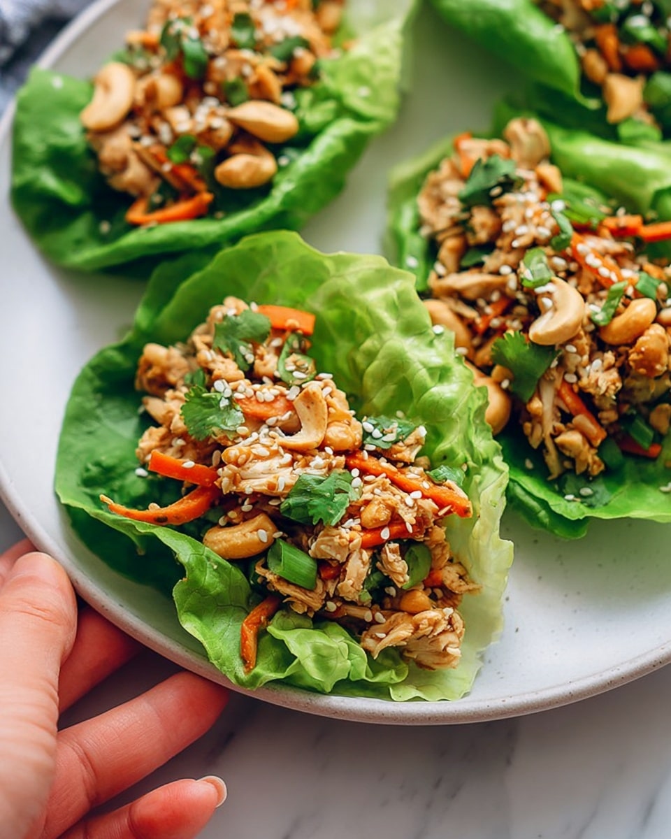 Three green lettuce leaves are placed on a white plate with a white marbled background. Each leaf acts as a wrap filled with chopped cooked chicken pieces mixed with small bright orange carrot bits, green onion slices, fresh cilantro leaves, and whole light tan cashew nuts. White sesame seeds are sprinkled on top, adding texture. A woman's hand is gently holding the edge of the plate. photo taken with an iphone --ar 4:5 --v 7