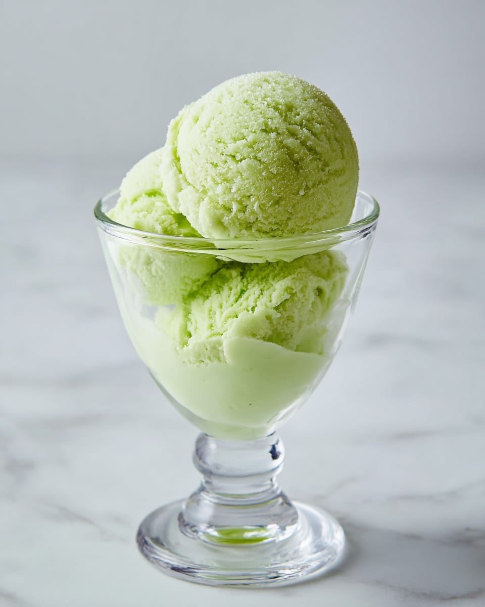A clear glass dessert cup holds two scoops of pale green ice cream. The bottom scoop fills the glass almost to the top, showing a smooth, slightly creamy texture with a soft, muted green color. Sitting on top is a second scoop with a rougher texture, showing small ice crystals, and a brighter, fresh light green shade. The glass stands on a thick, clear base and is set on a white marbled surface, making the green ice cream stand out clearly. photo taken with an iphone --ar 4:5 --v 7