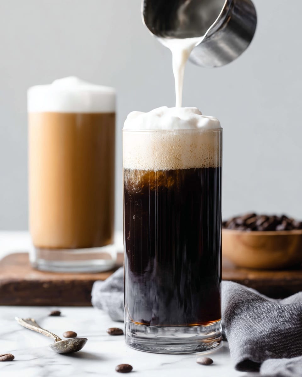 The image shows a tall, clear cylindrical glass filled with dark black coffee, with creamy white foam being poured from a silver cup above, creating a soft thick layer slowly covering the top of the coffee. Behind this glass is another tall glass filled with a lighter brown coffee topped with thick white foam. The scene is set on a white marbled surface with a grey cloth and a small spoon in the foreground. There is also a small wooden bowl filled with dark coffee beans in the background, adding contrast and texture. photo taken with an iphone --ar 4:5 --v 7